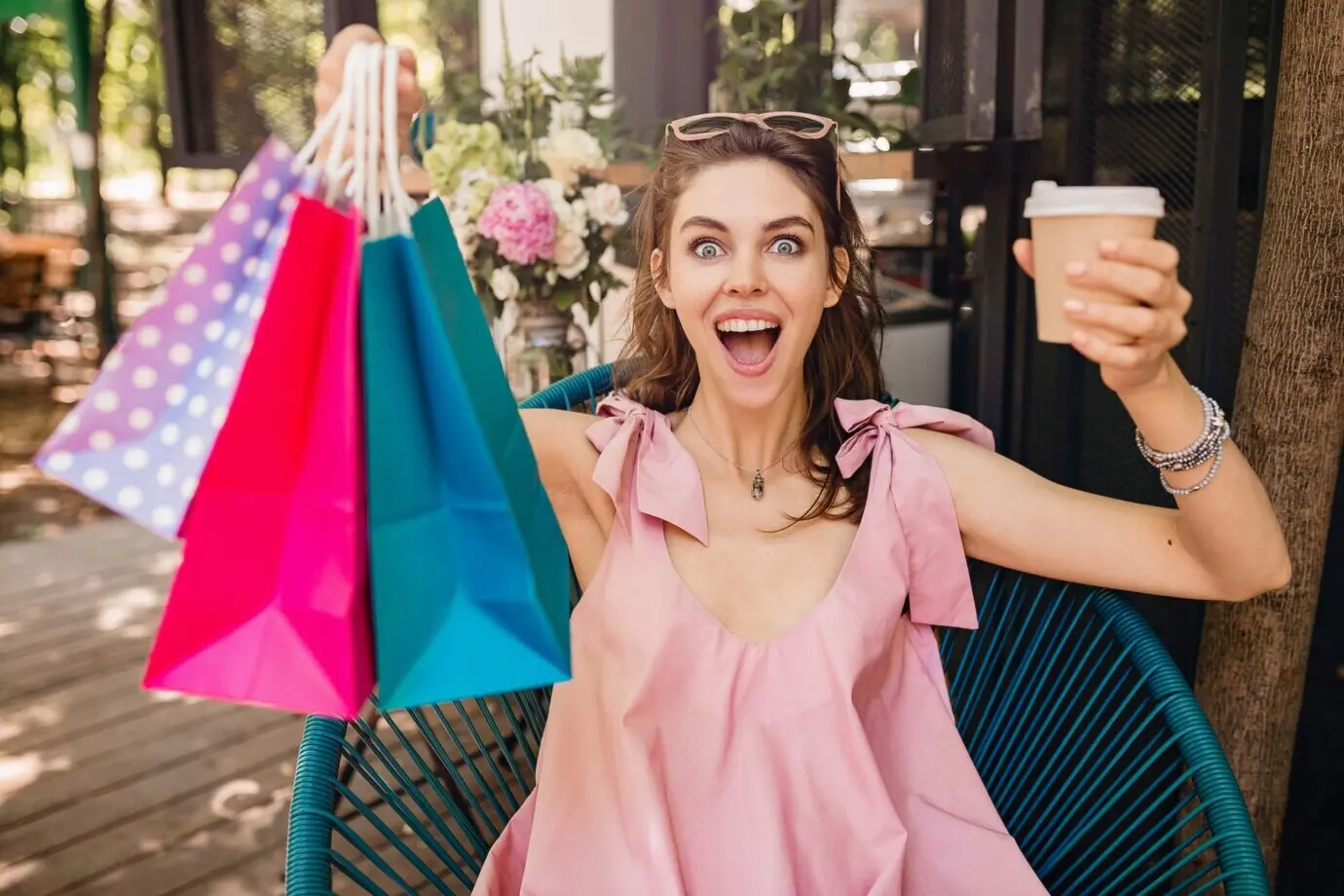 Retrato de una mujer joven, bonita, sonriente y feliz, con expresión facial emocionada, sentada en una cafetería con bolsas de compras y bebiendo café, con un atuendo veraniego de moda y de tendencia: un vestido rosa de algodón.