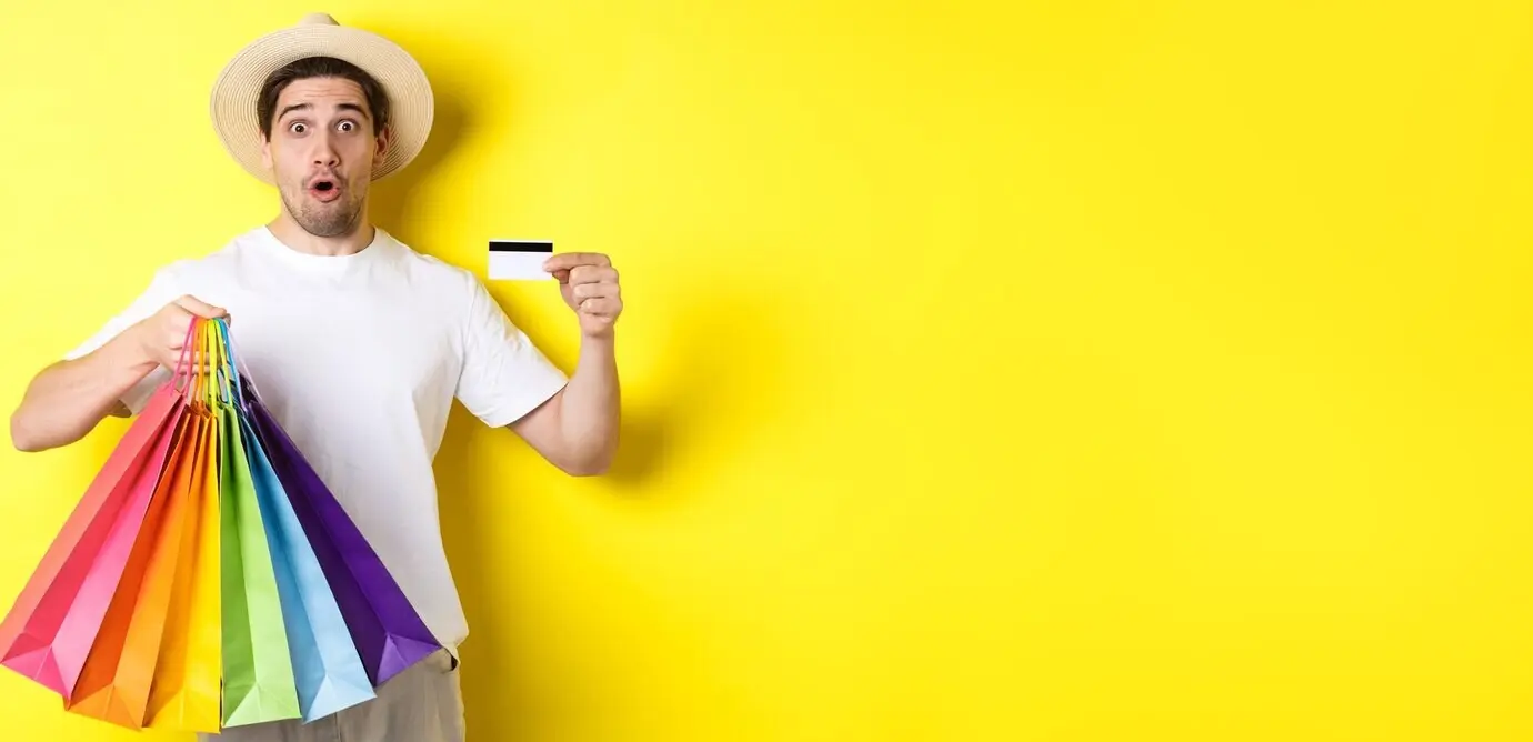 Hombre impresionado mostrando bolsas de la compra con productos y una tarjeta de crédito, de pie sobre un fondo amarillo.